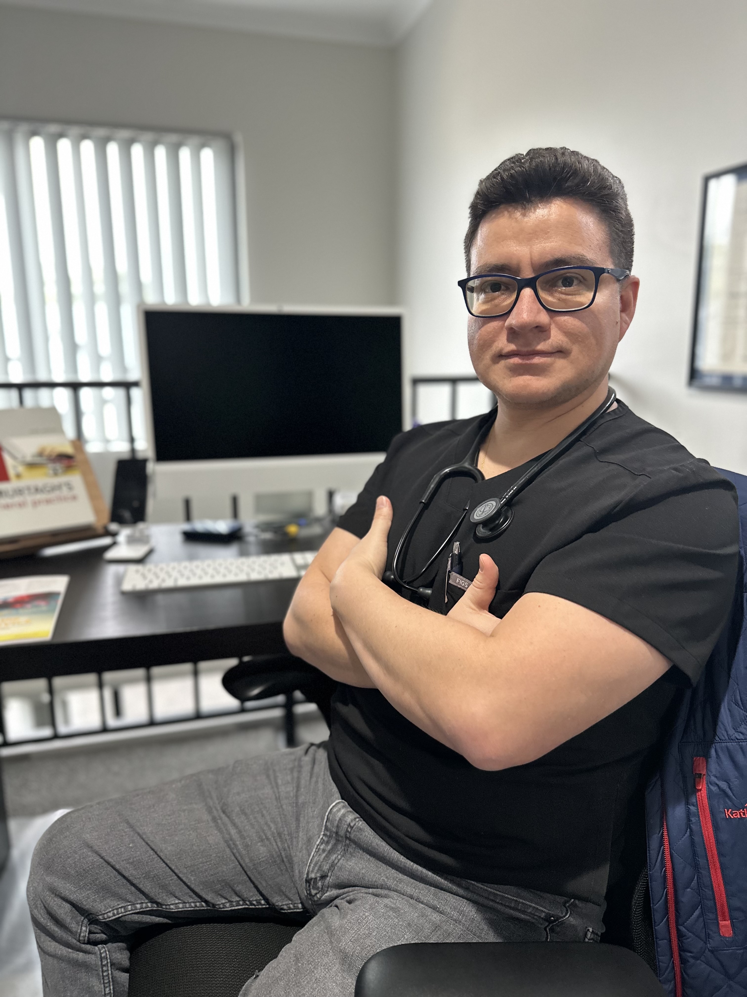 Dr. Sebastian Calvache Rubio sitting at a desk in a medical office, wearing a stethoscope and scrubs
